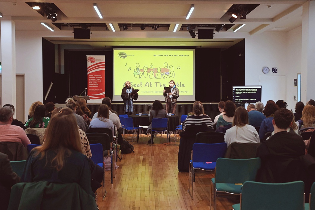 Audience seated in chairs in a conference room, facing two hosts standing at the front of the room in front of a projector screen, reading from clipboards.
