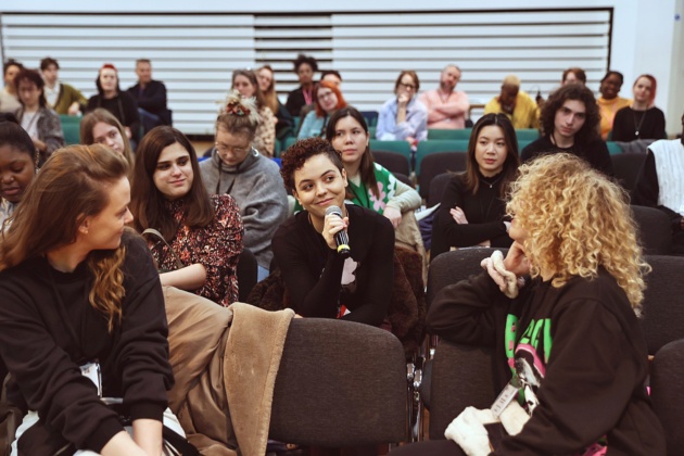 People sitting in chairs, having a discussion in a social setting. One in the centre holds a microphone and is smiling.