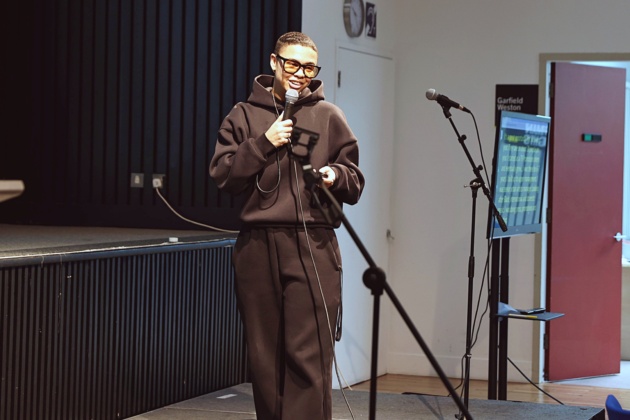 A person standing at a podium with a microphone smiles and addresses an audience that we can't see.