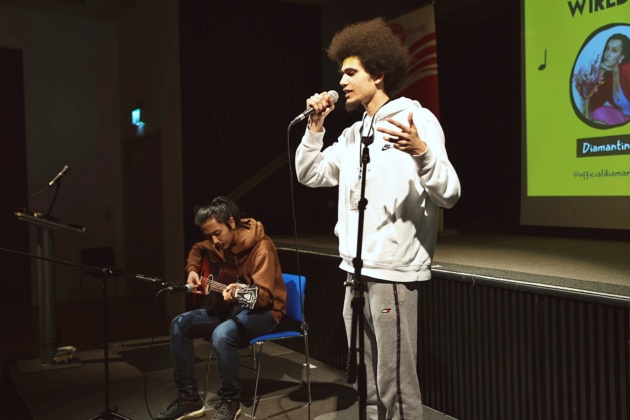 Young man stands rapping into a microphone. Another young man is seated beside him playing the guitar.